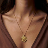 Gold necklace with a circular pendant worn by a person in a brown top.